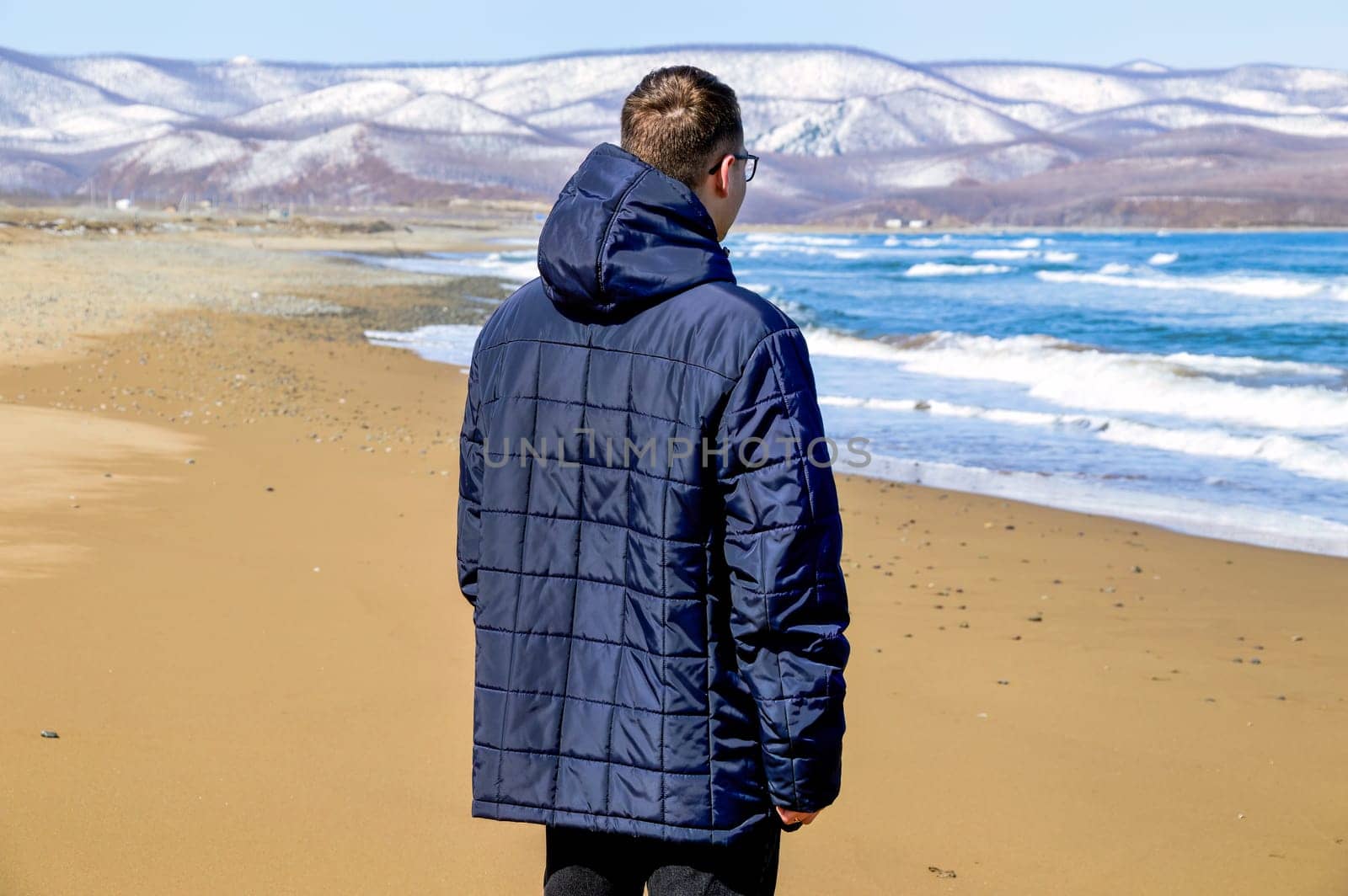 Brunette young man in jacket walking on the beach close to snowy mountains and Japanese sea. Solo traveller.