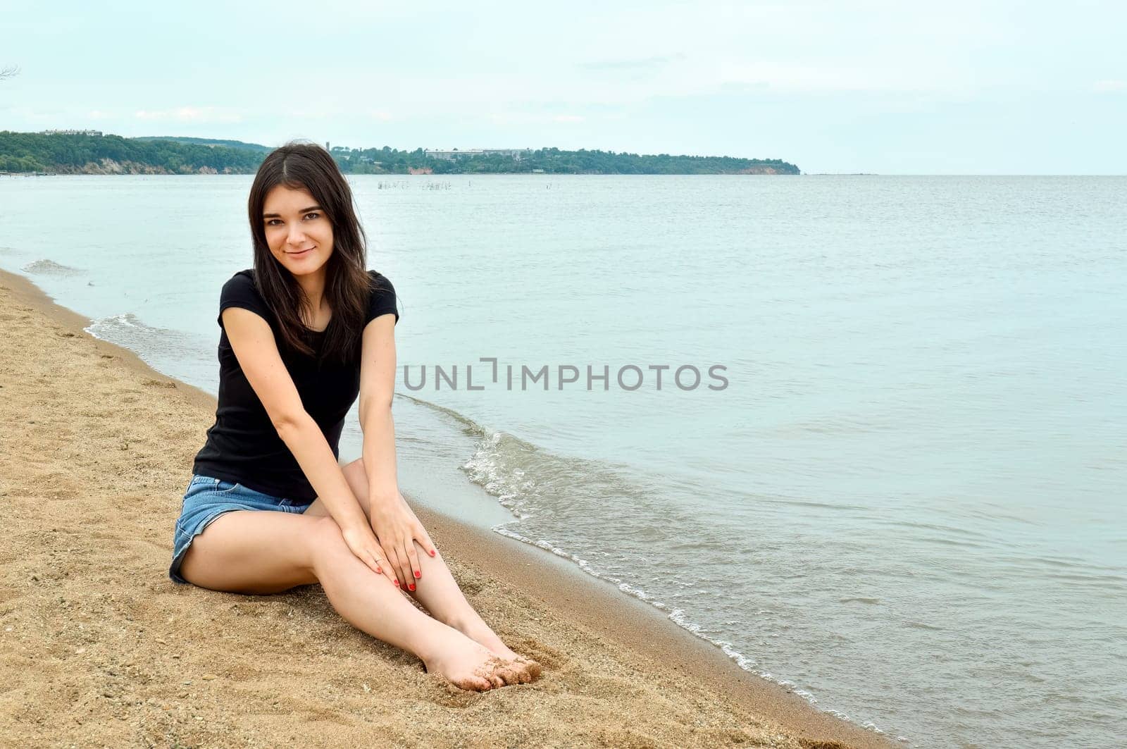 Attractive brunette girl sitting near water on a cloudy summer day. Lake Khanka, Russia.