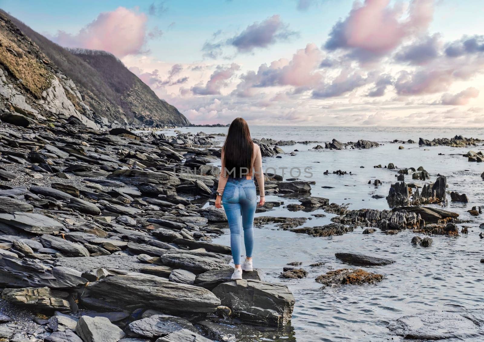 Young slender woman walking at the beach with black stones and looking at the sunset