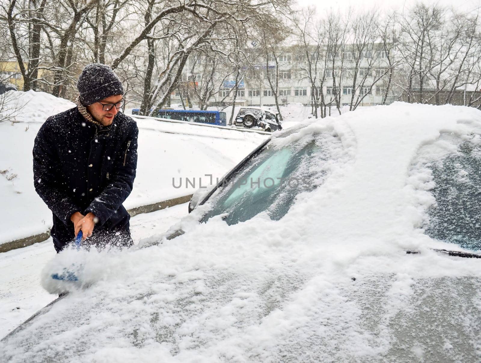 Young man sweeps snow from the car on a snowy day in winter