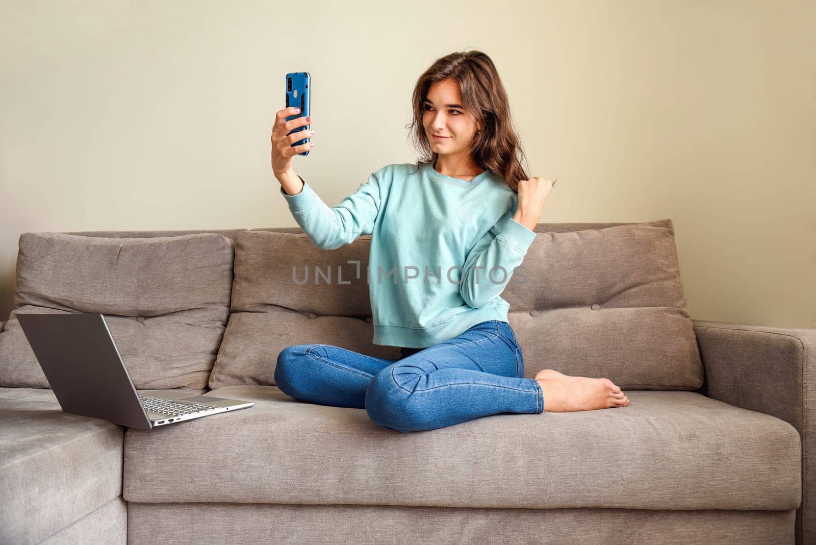 Young smiling woman taking a selfie for her profile. She sitting on  couch close to laptop at home. lockdown and online dating concept