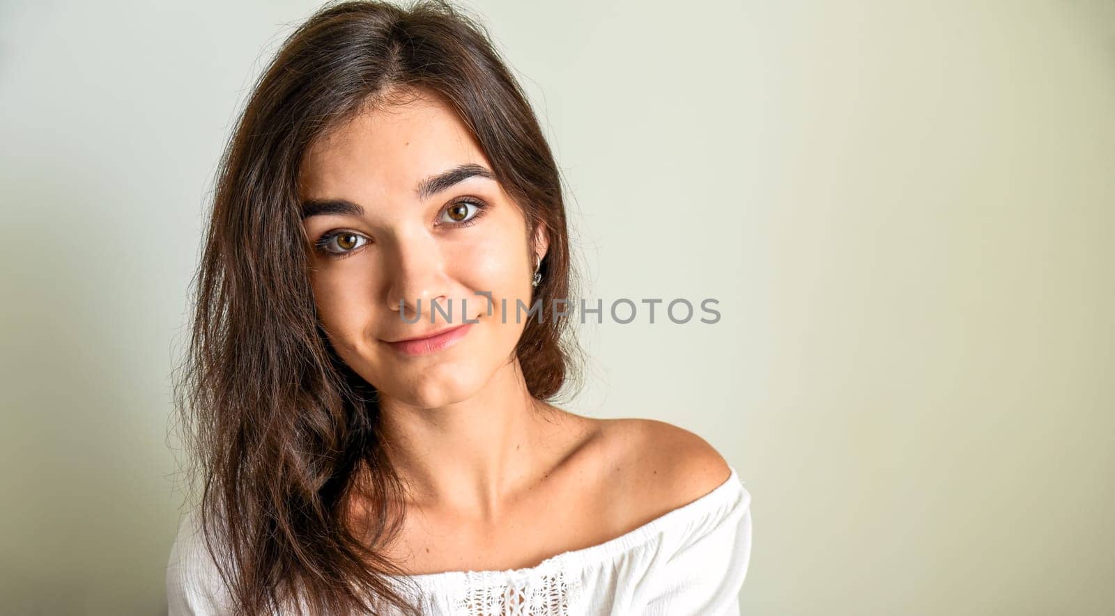Portrait of young 25 y.o. brunette Russian woman with long hair. In white open shoulder top and looking into the camera on light gray background. Minimum makeup, natural hairstyle. copy space