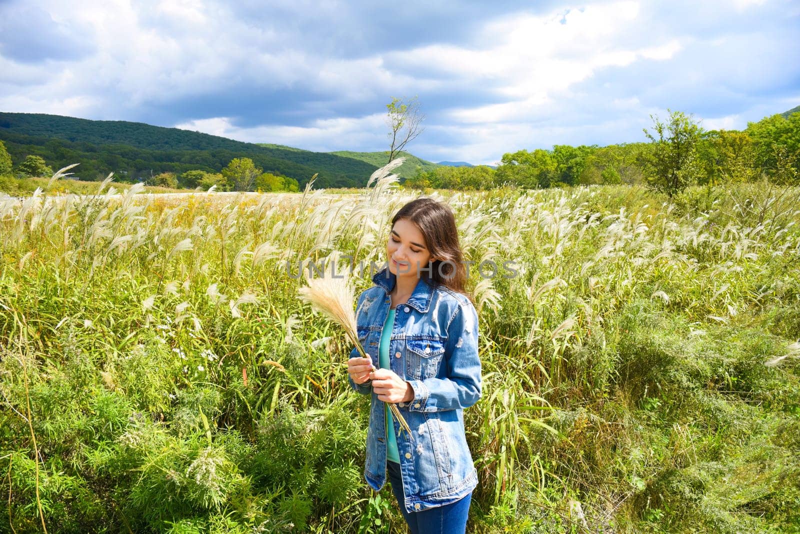 Young smiling woman holding bouquet of Miscanthus sinensis (Chinese silver grass) in her hands and looking at it against the background of a field. Autumn in Russia. Copy space