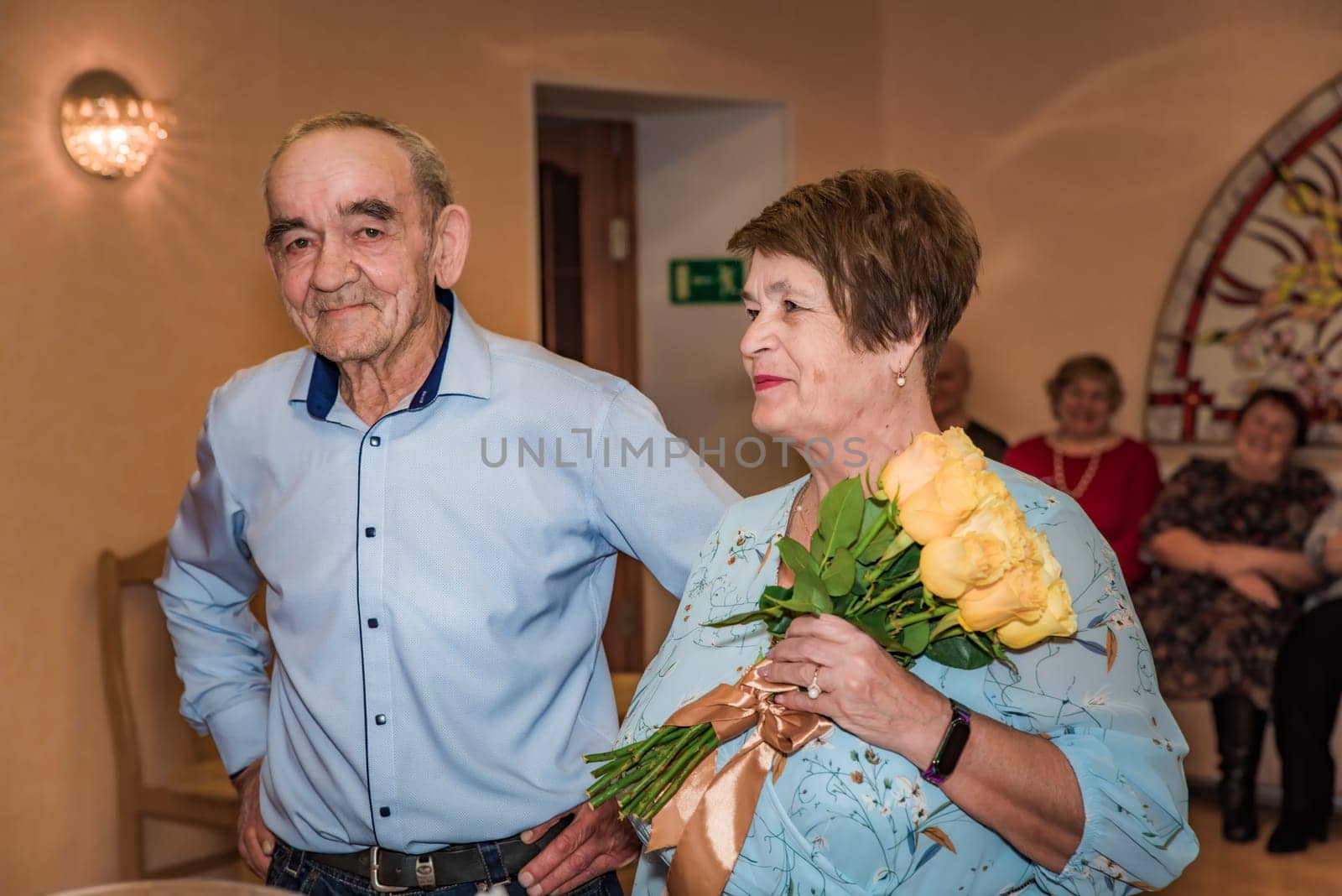 Russian elderly couple, both in their 70s, celebrates their wedding at a registry office. The bride holds a bouquet of yellow roses while friends sit in the background. Both are smiling and elegantly dressed