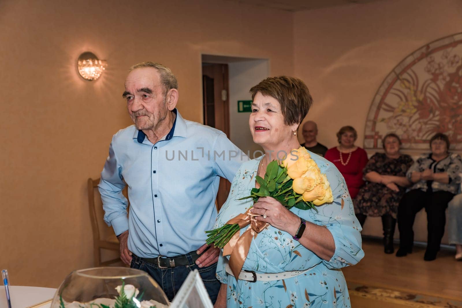 A couple celebrates their 50th wedding anniversary (golden wedding) in the registry office. The wife smiles while holding a bouquet, and their friends are seen in the background, all dressed up
