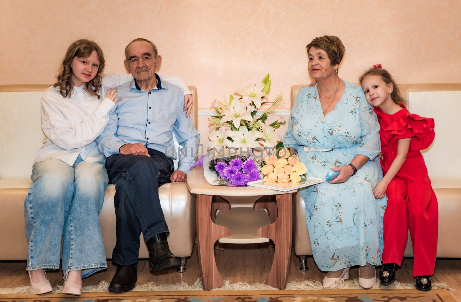 Two granddaughters sit with their Russian grandparents, celebrating their 50th golden wedding anniversary. The family is seated on a sofa, surrounded by bouquets of flowers