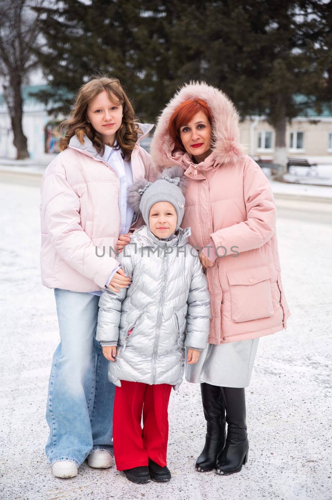 Russian single mother with her teenage and 8-year-old daughters standing in a snowy city. All wear warm winter outfits, including pink and silver jackets. Stylish family winter fashion outdoors