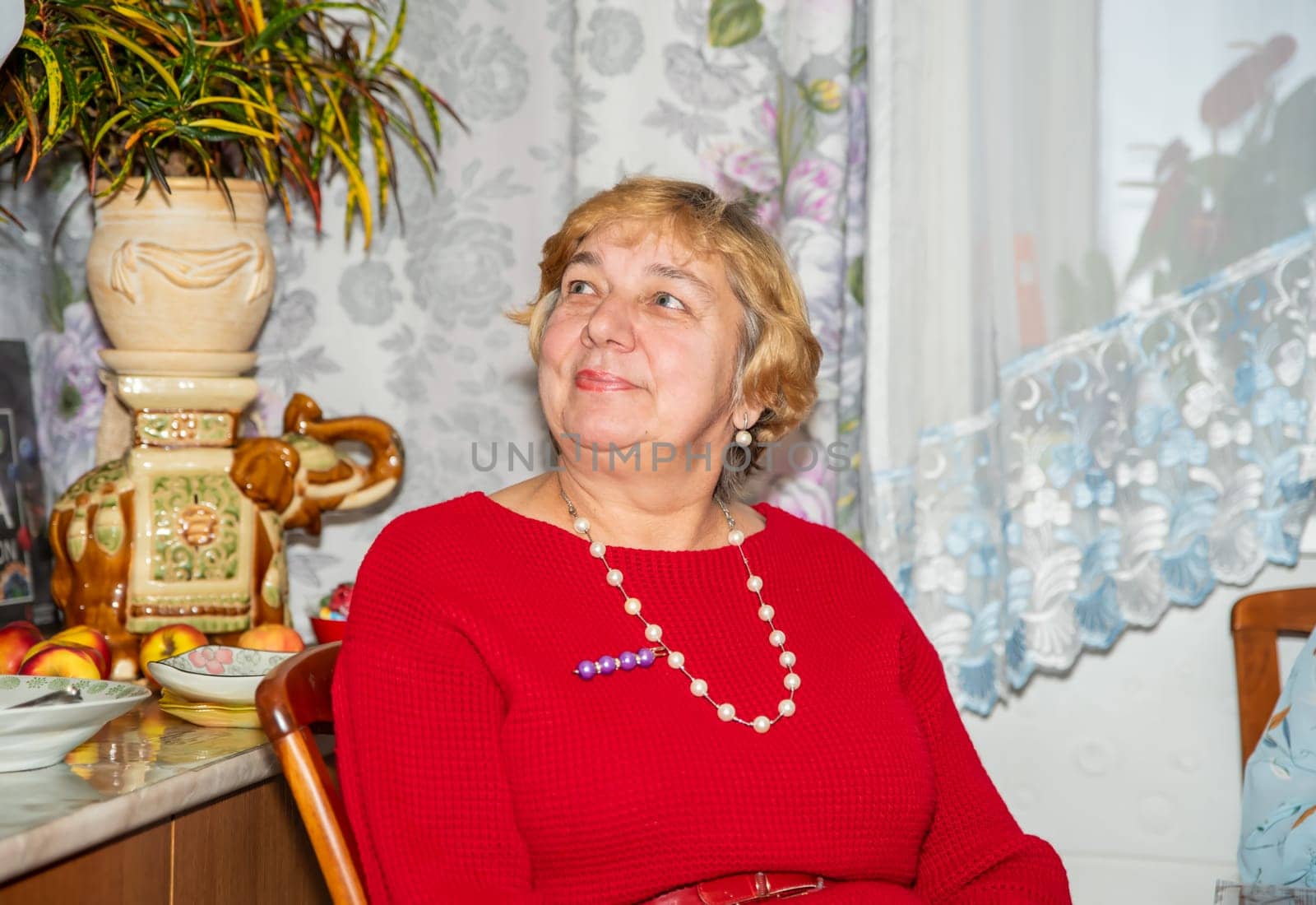 A 60-year-old Russian woman in a red warm dress with beads and short hair sits on a chair at home, looking to the left, enjoying her time as a guest in a cozy indoor setting