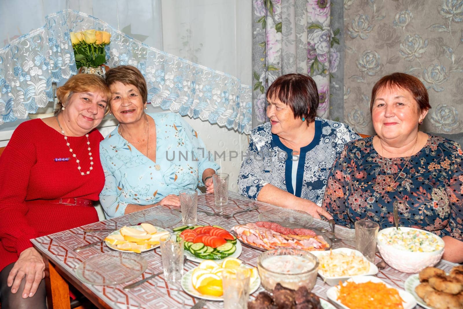Four Russian friends, two in their 60s and two in their 70s, celebrate a special occasion together. They smile and pose for the camera at the dinner table, enjoying the moment with warmth and joy