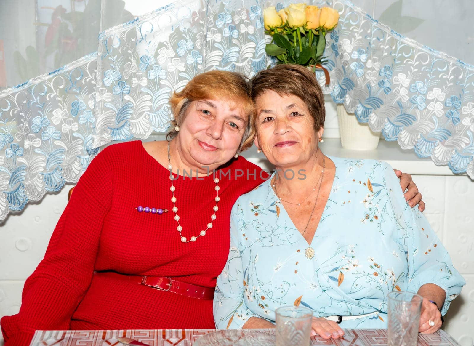 Two cheerful Russian women, aged 60 and 71, smile and embrace at home, celebrating their lifelong friendship. A warm moment of togetherness, perfect for themes of senior friendship and positive aging.