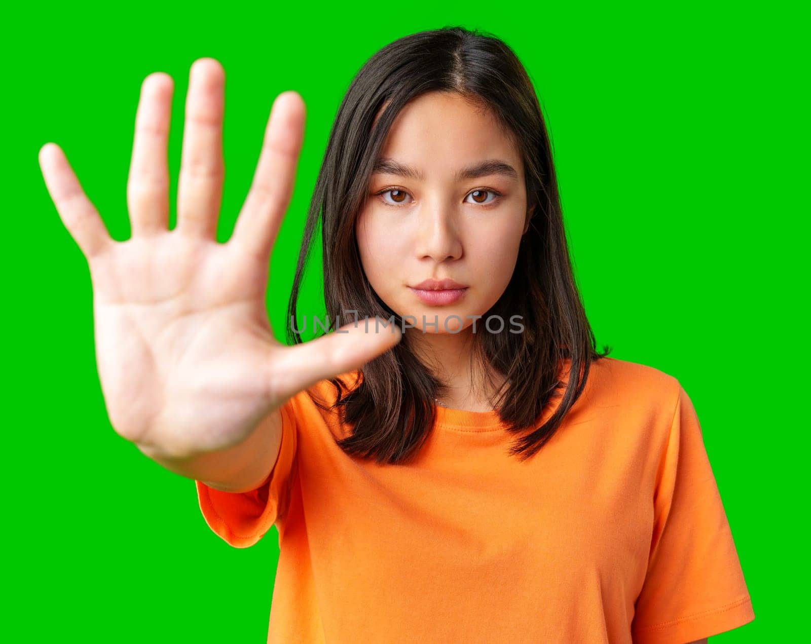 A young woman stands confidently with her hand raised in a stop gesture. She is wearing an orange t-shirt and has dark hair. The vibrant green background enhances the focus on her expression.