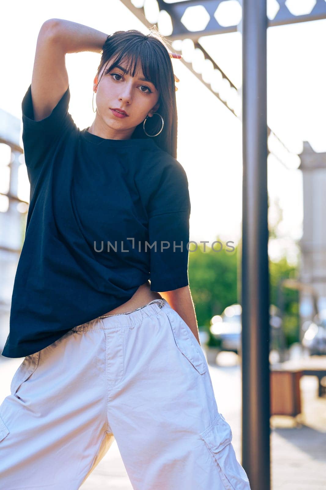 A young woman showcases her style in a relaxed black top and light pants while posing in an urban setting. The sun casts a warm glow, highlighting her confident demeanor.