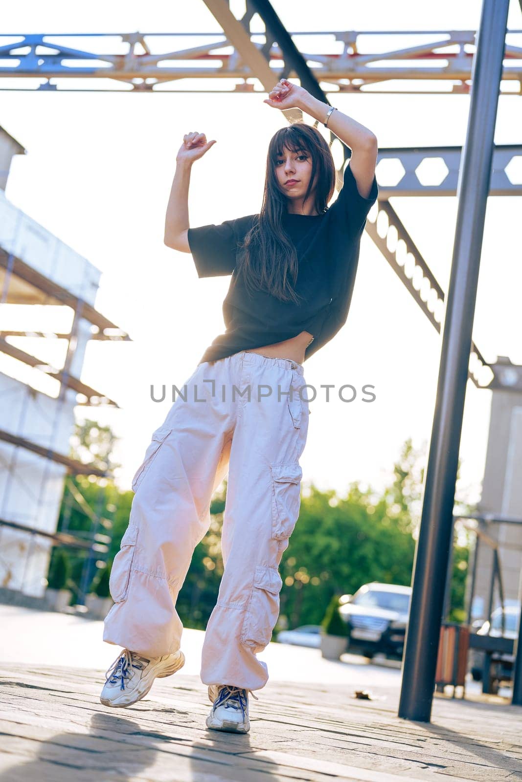 A young woman with long hair is dancing joyfully outdoors, bathed in golden sunlight. She wears a loose black top and light-colored cargo pants, showcasing an energetic vibe amidst an urban backdrop.