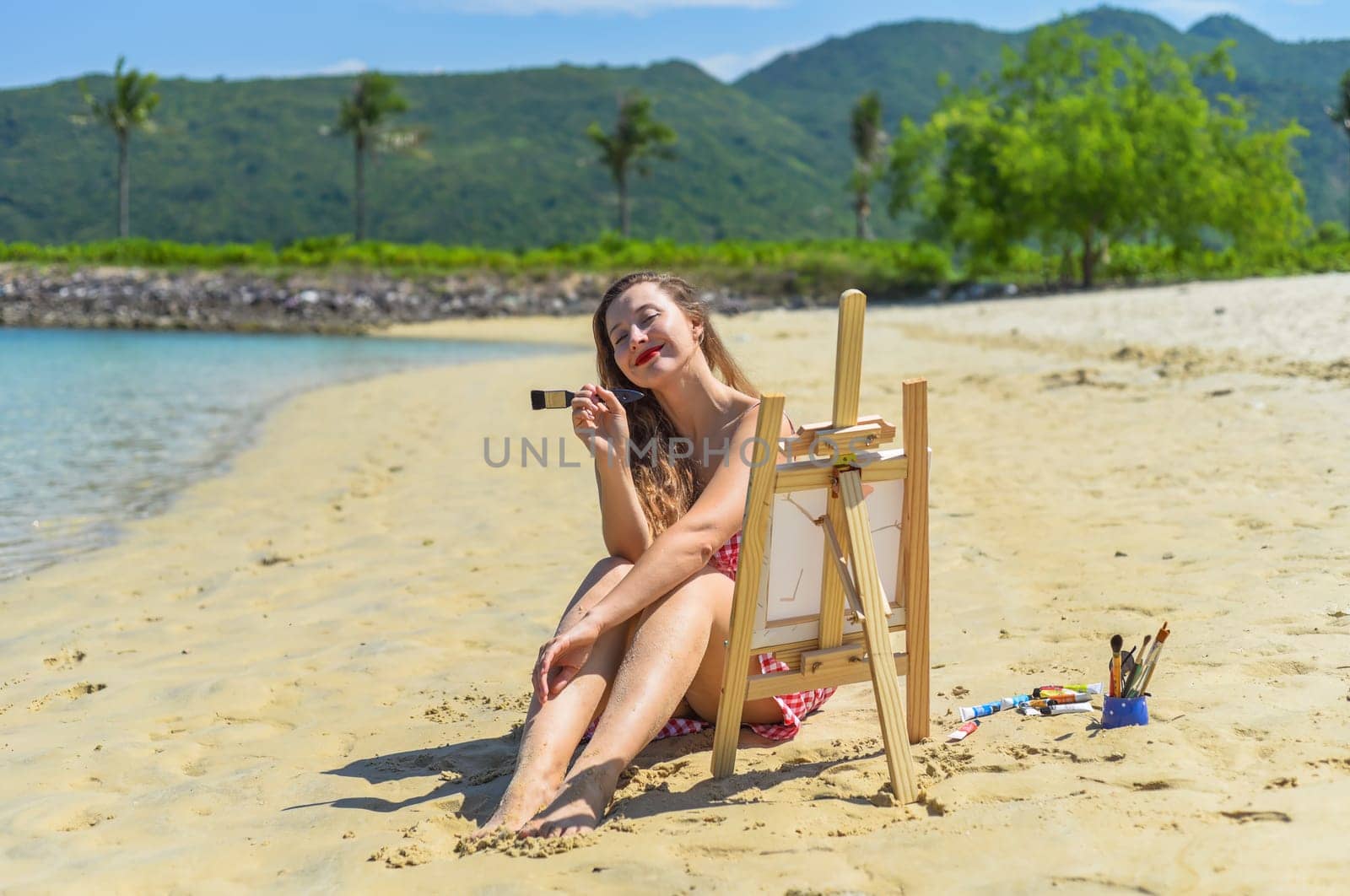 Young artist woman sitting at the beach with a jar with paint brushes and easel close to sea and palms in summer
