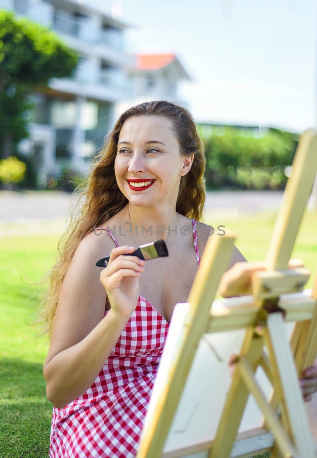 Young artist woman holding a jar with paint brushes and easel close to sea and palms in summer