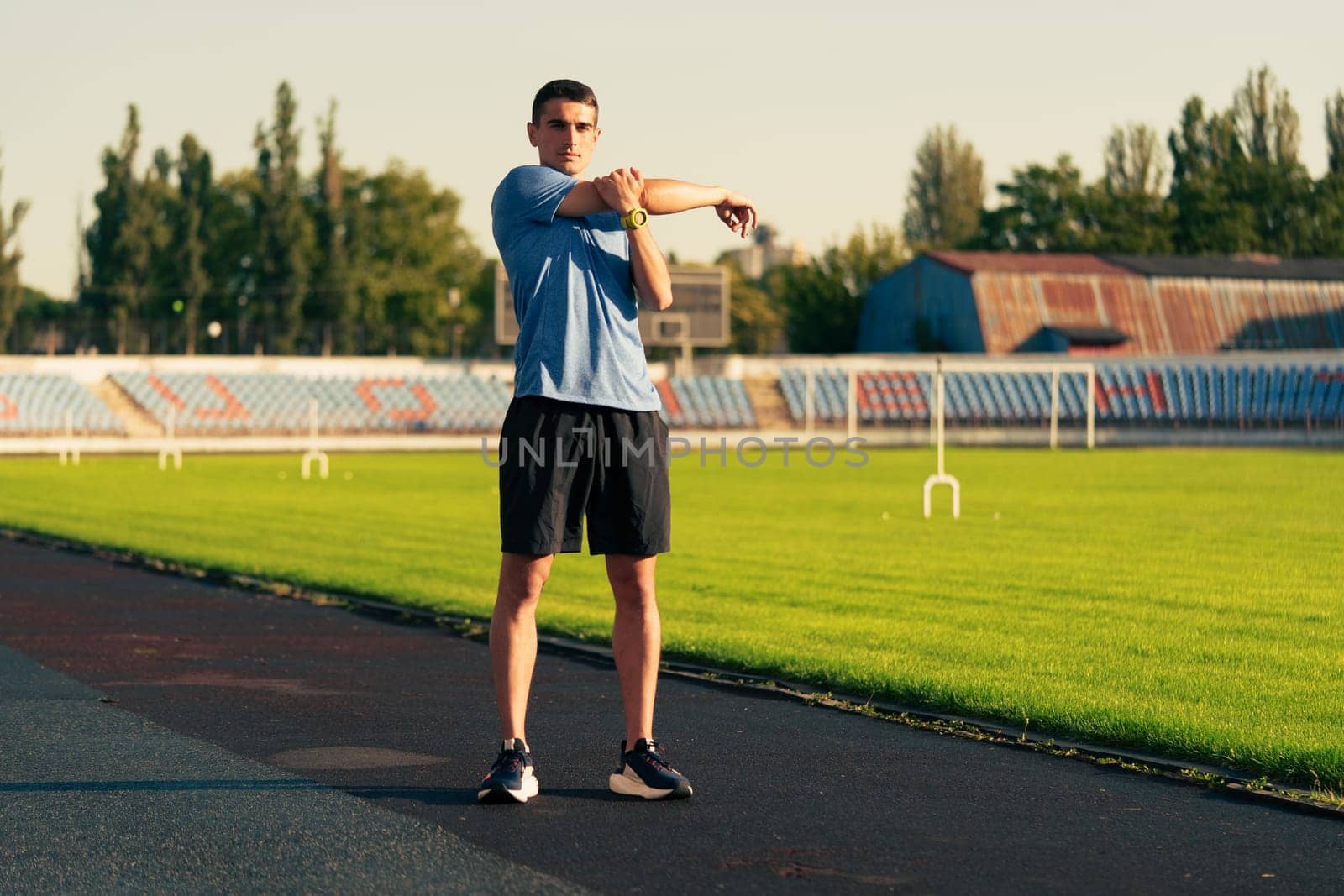 A young man in athletic wear stretches his arms on a running track surrounded by green grass and stadium seating. The sun casts a warm glow while he prepares for his workout.