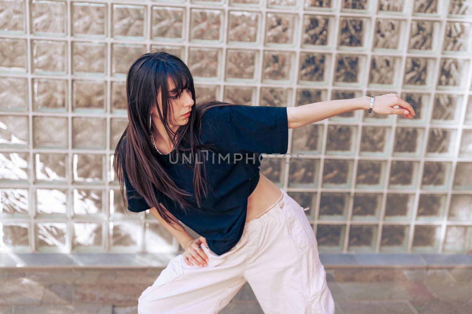 A young woman performs dynamic dance moves in an urban setting, wearing casual attire. Sunlight highlights her energetic expression and stylish form, creating a lively atmosphere.