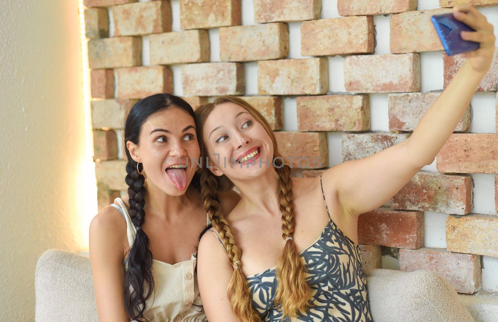 Ukrainian woman (brunette) and Russian woman (blonde) portrait together with braids posing and taking a selfie, showing tongue