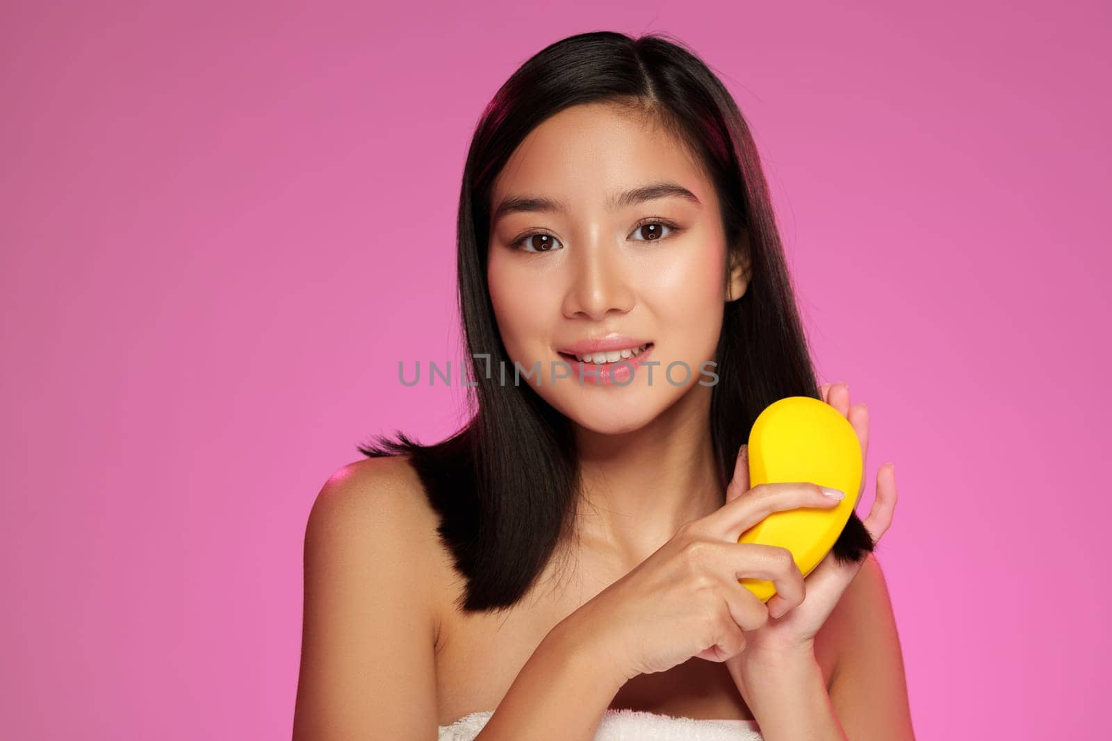 A woman with straight black hair holds a yellow skincare tool in her hand while smiling. She poses against a bright pink background, highlighting beauty and skincare products.
