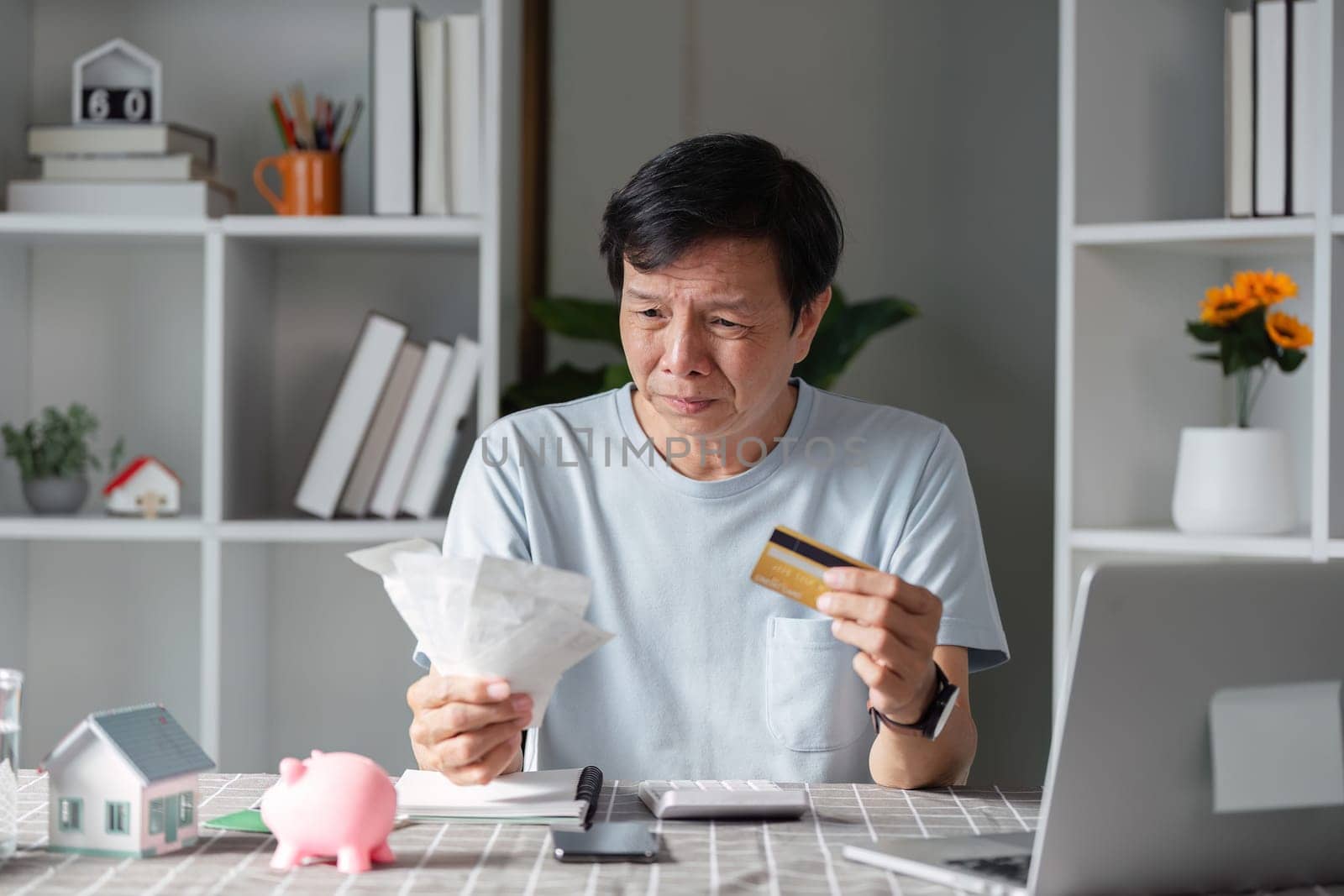 A concerned man evaluating his financial situation, holding bills and a credit card, expressing worry in his home office filled with paperwork.
