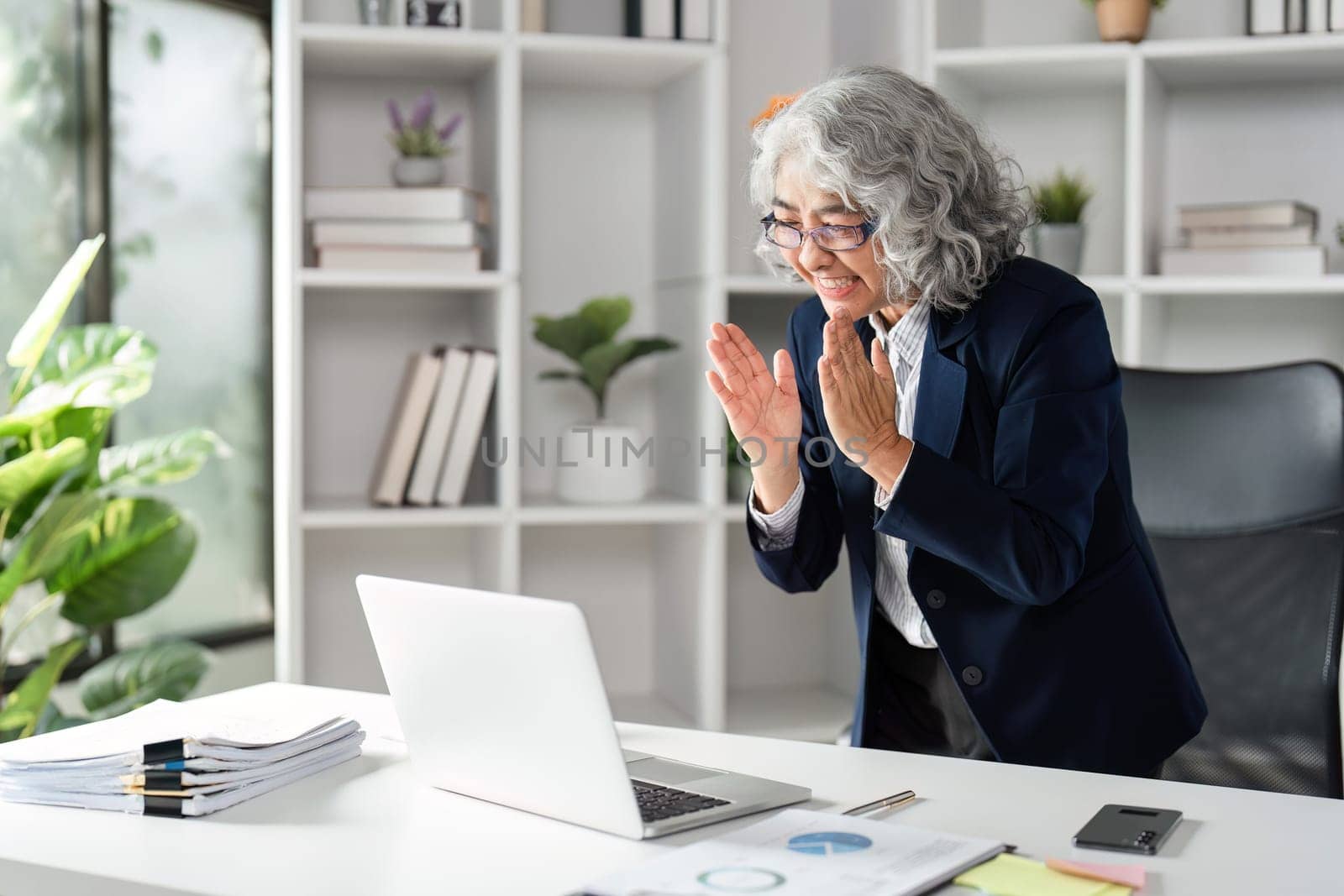 An elderly businesswoman shows her excitement and happiness for a recent accomplishment, interacting with her laptop in a modern office setting.
