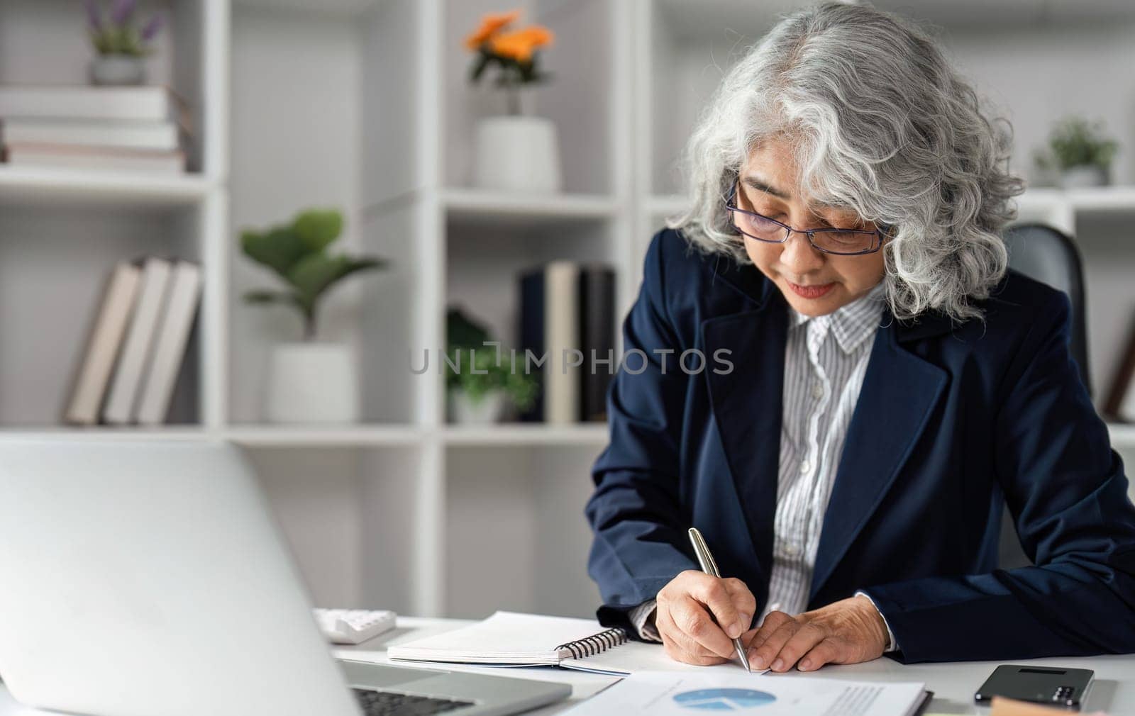 An elderly business woman is focused as she takes notes and analyzes data charts at her contemporary desk, highlighting her dedication and professionalism.
