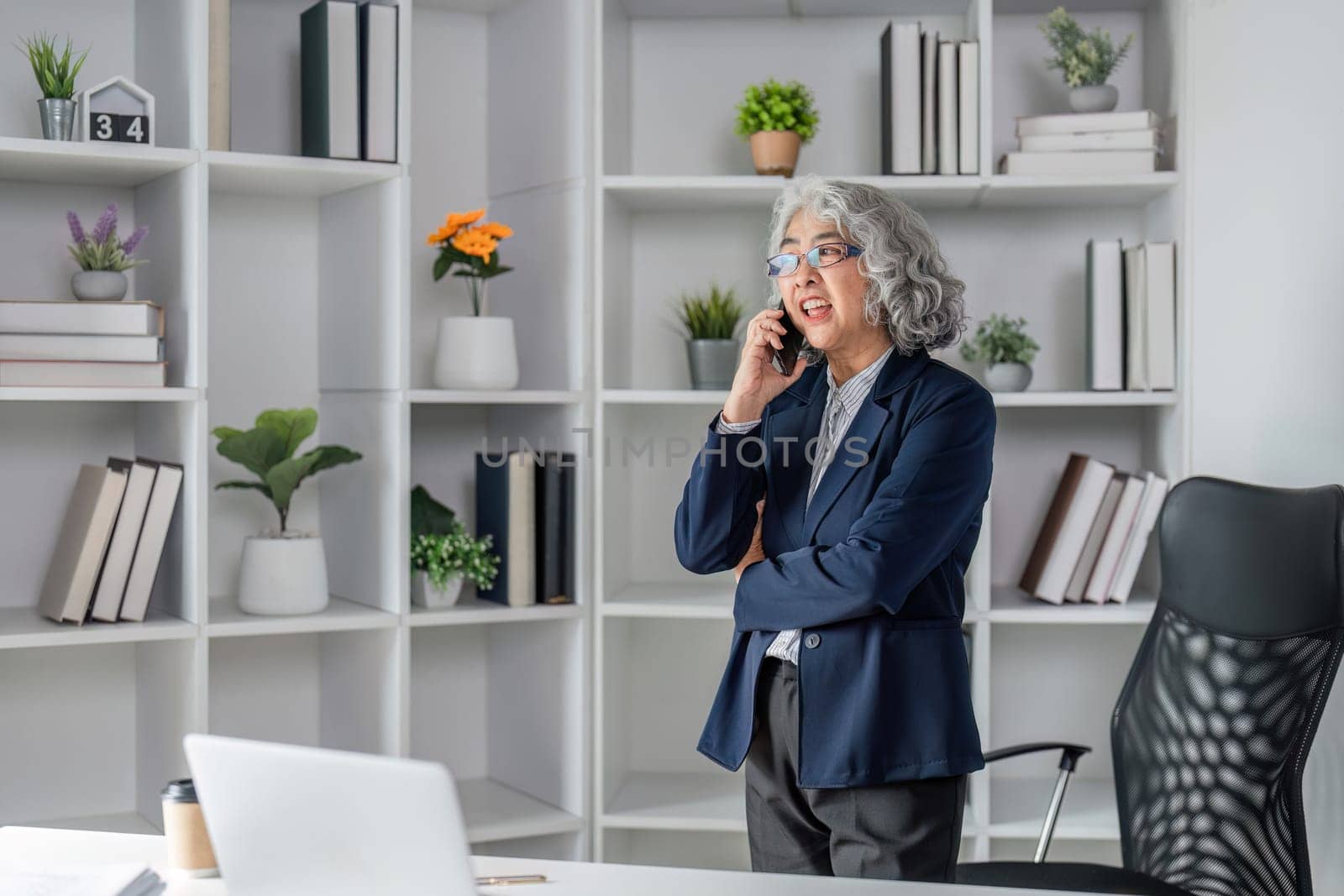 An experienced senior business executive engaged in a strategic phone conversation, standing confidently in a well-organized modern office with shelves and plants.