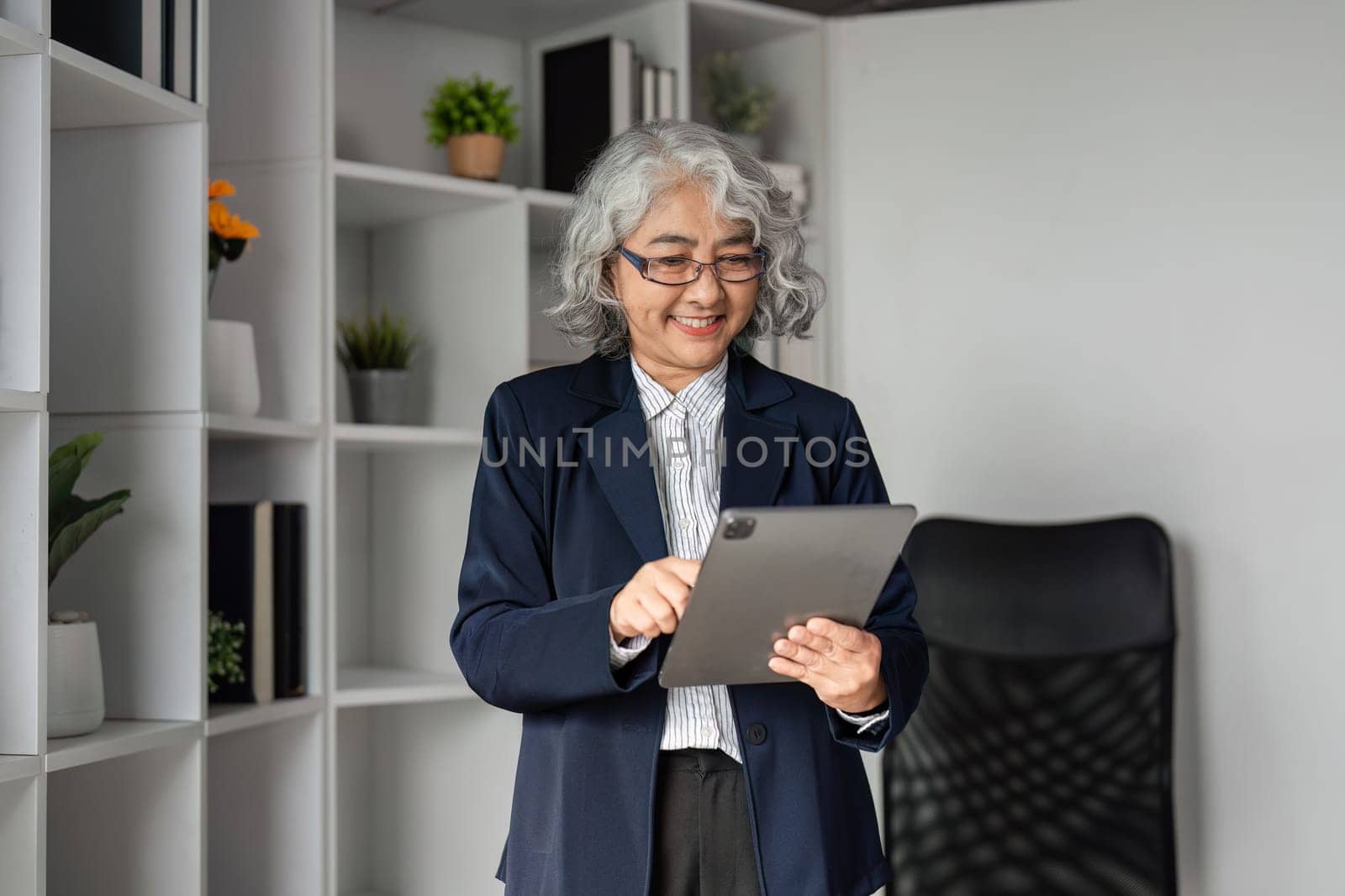 A senior business executive with gray hair happily interacts with a tablet, standing confidently in a contemporary office space filled with plants and organized shelving.