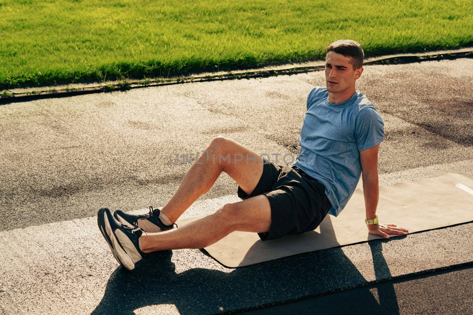 A man performs a workout on a mat in an outdoor park, showcasing focus and determination. The bright daytime setting highlights the lush green grass surrounding him.
