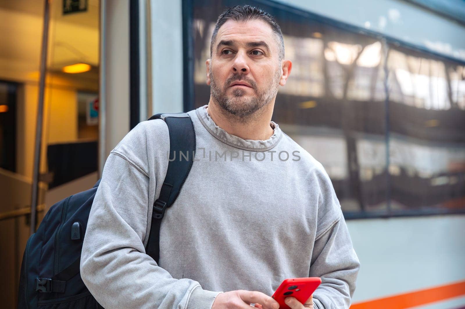 Serious man wearing a gray sweatshirt holding a red smartphone while looking around at a train station. His expression conveys anticipation, travel, and modern connectivity.