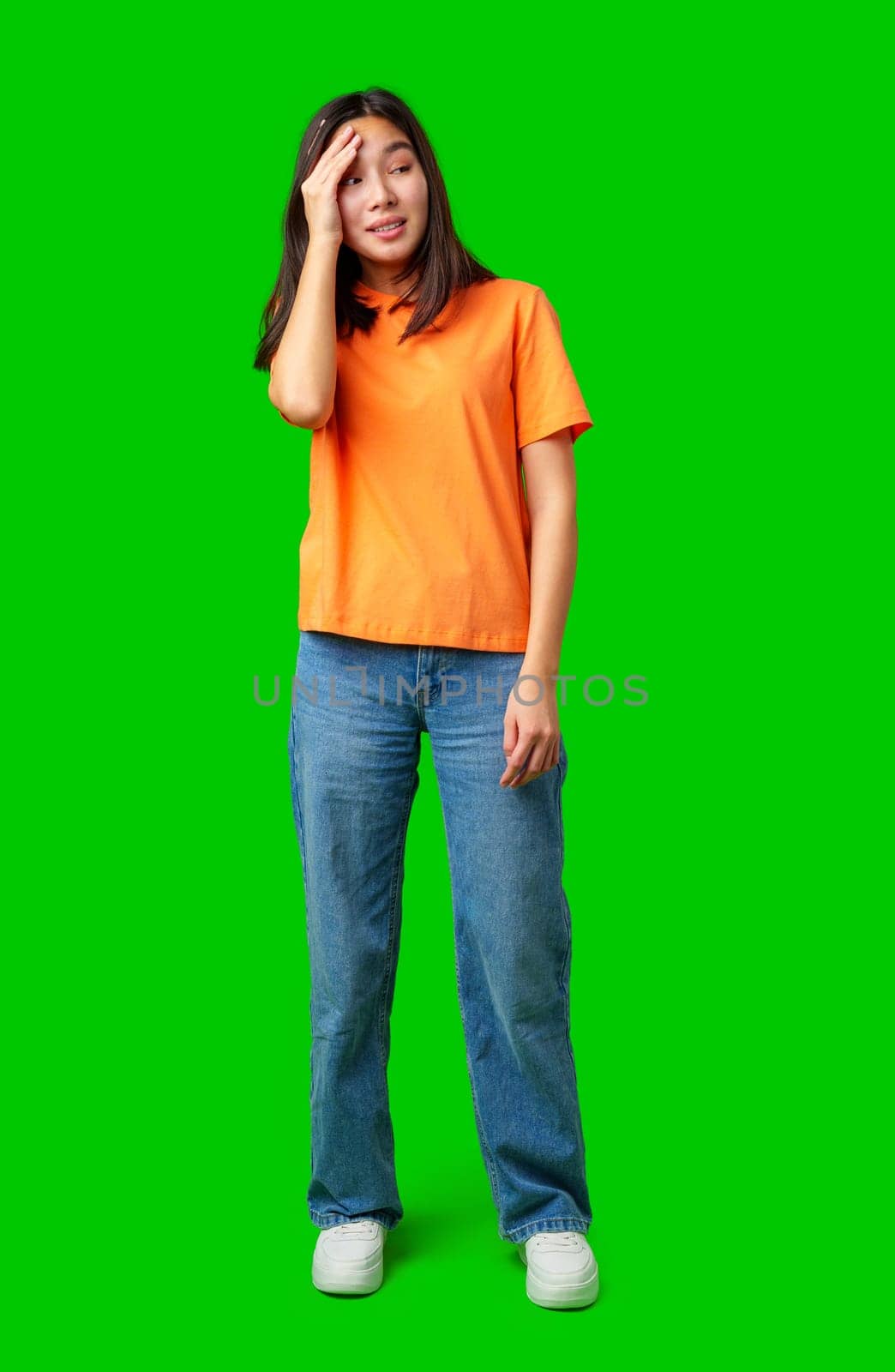 A young woman stands in a bright orange shirt and blue jeans, showing a confused expression. She touches her head, surrounded by a vibrant green backdrop, indicating a playful mood.