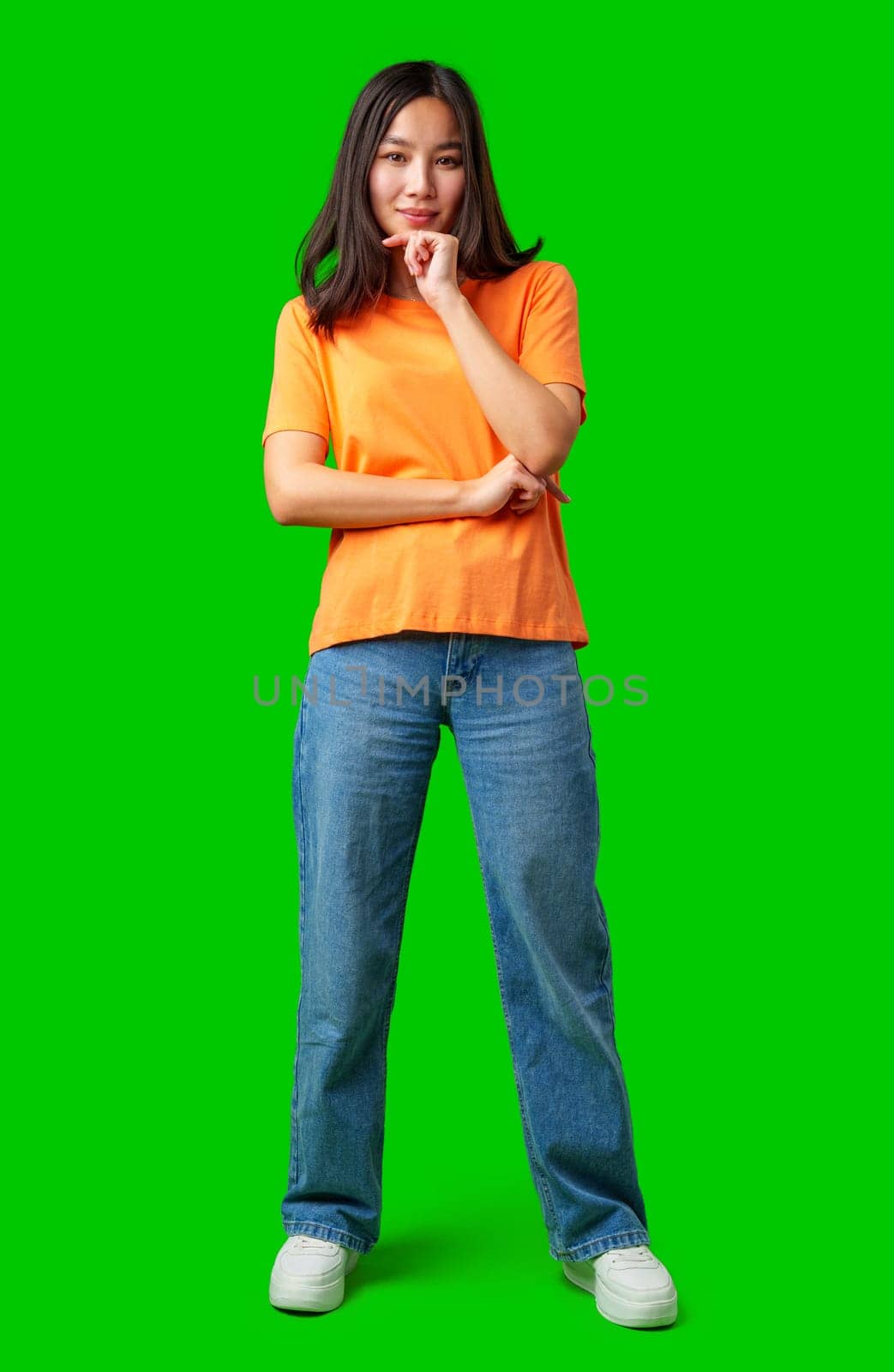 A young woman stands with her arms crossed, wearing a bright orange shirt and blue jeans. She has a thoughtful expression while against a vibrant green background.