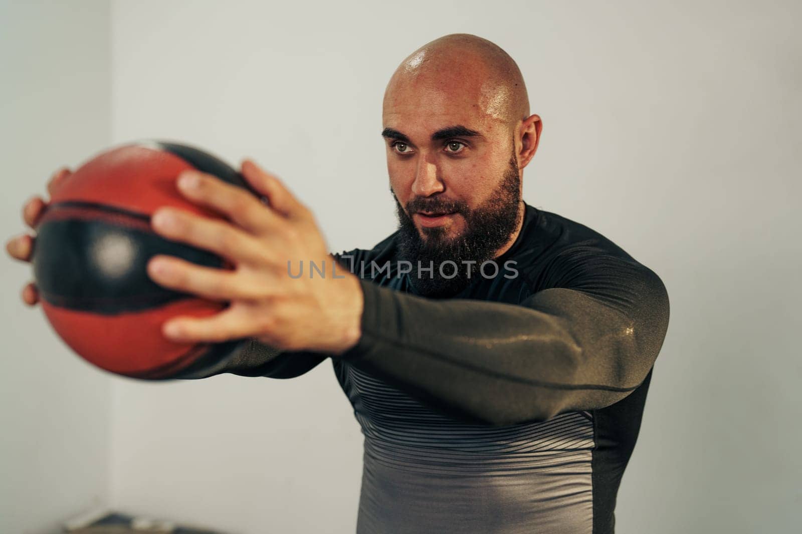 A man focuses on his workout routine by performing strength training with a medicine ball in a well-lit gym. He displays concentration and determination while exercising.