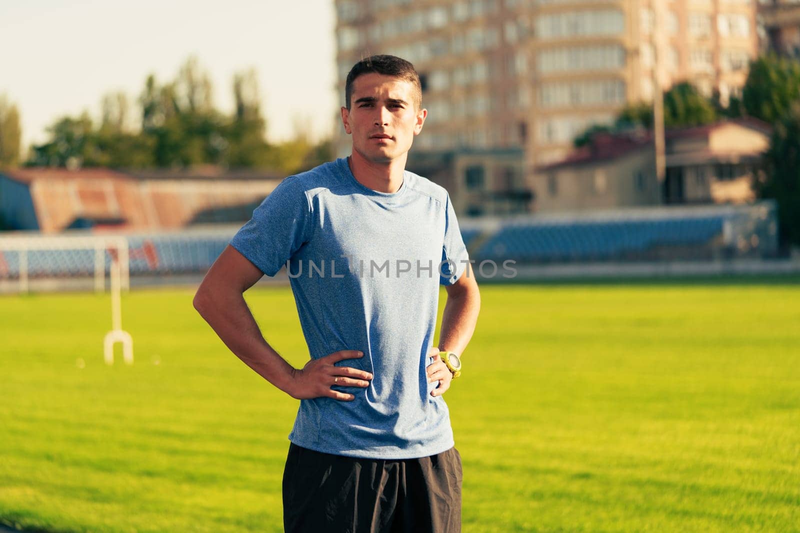 A young male athlete stands on the track field with hands on hips, gazing confidently ahead. The sun shines brightly, illuminating the green grass and nearby buildings.