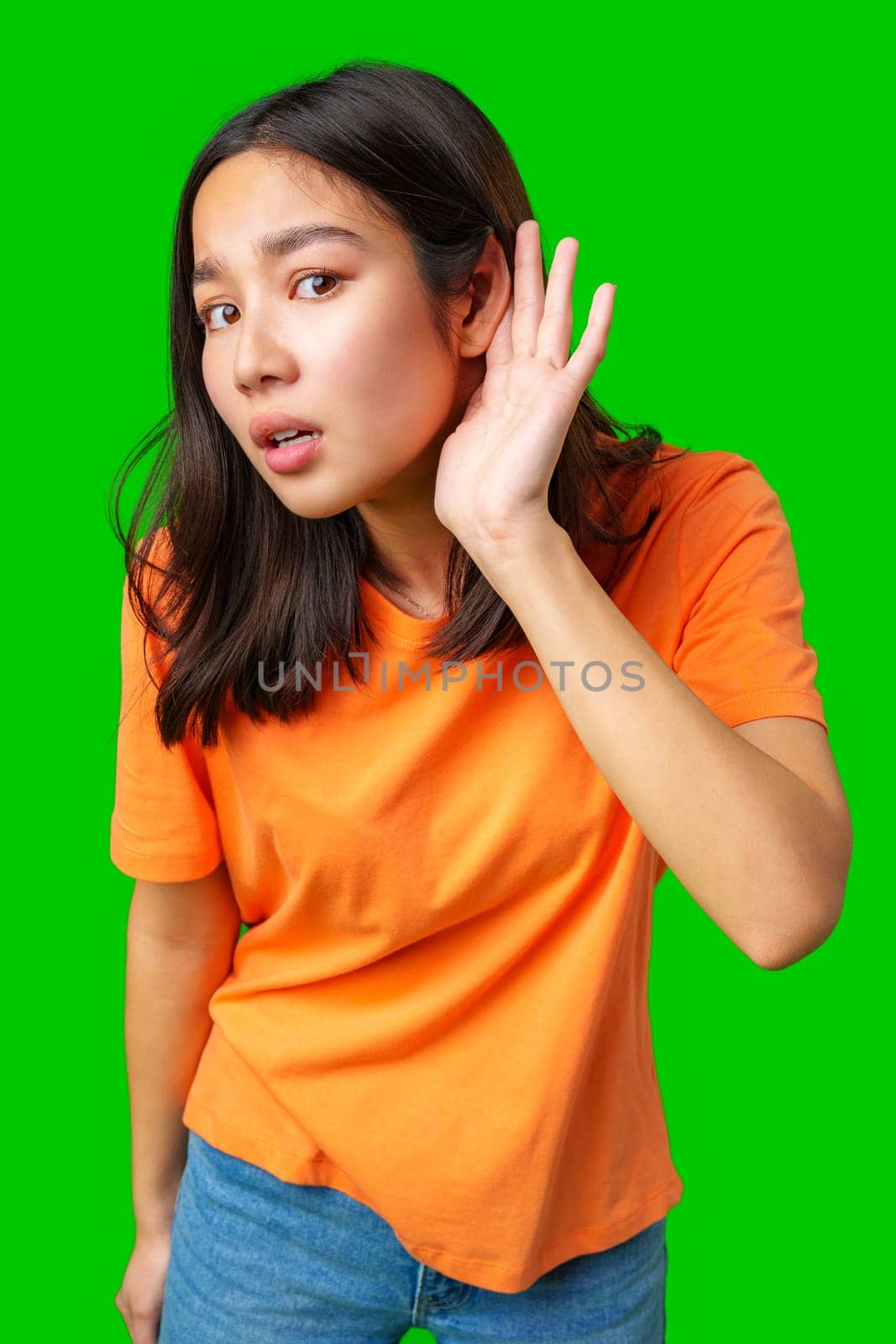 A young woman with long black hair, wearing an orange shirt, leans in with a curious expression as she listens attentively. The bright green background enhances her engagement with the viewer.
