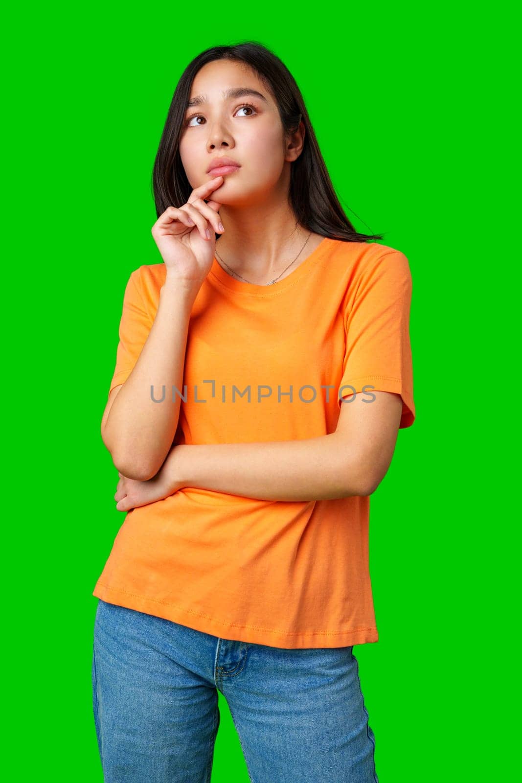 A woman wearing an orange t-shirt stands with her arms crossed, looking thoughtfully to the side. The bright green backdrop creates a lively contrast, adding energy to the moment.
