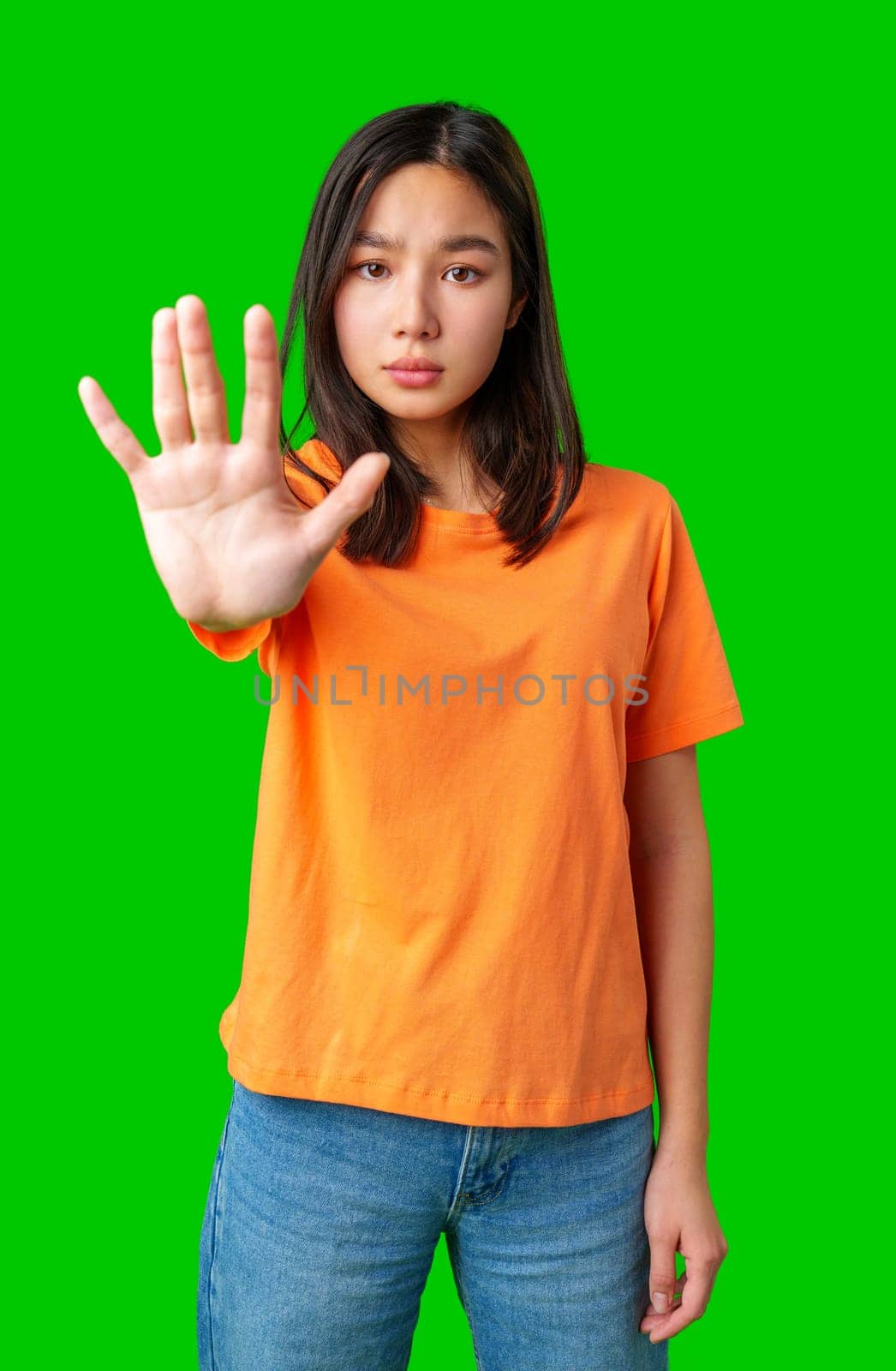 A young woman stands confidently in front of a bright green background, raising her hand in a stop gesture. She wears a casual orange shirt and jeans, expressing strong determination.