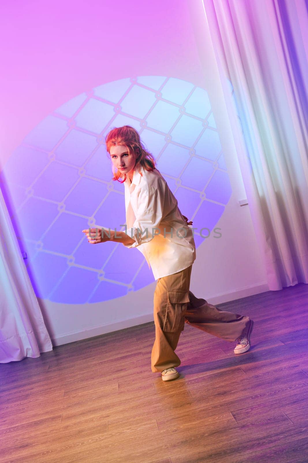 A contemporary dancer showcases their unique style in a vibrant studio, illuminated by colorful lights and a striking geometric projection during an evening performance.