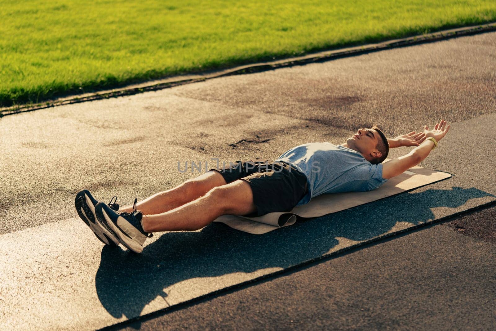 A person lies on a yoga mat in a park, stretching with arms extended. The bright sunlight reflects on the green grass, creating a serene outdoor exercise environment.