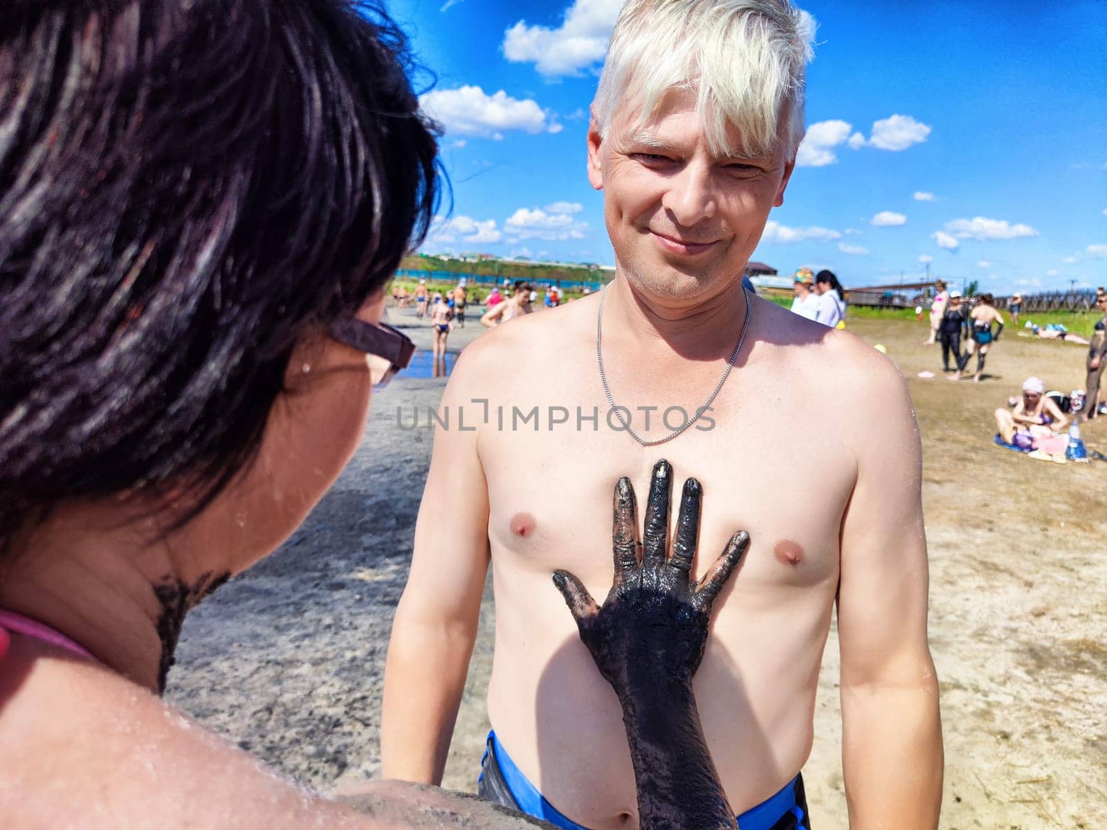 A middle-aged couple shares a fun moment at the beach, with the woman playfully applying mud to the man's chest. The vibrant blue sky and sandy surroundings enhance their joyful experience.