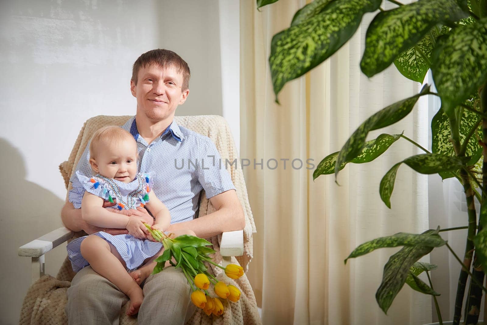 A joyful father sits in an armchair holding his smiling baby daughter, who is wrapped in his embrace while proudly clutching a bouquet of yellow tulips.
