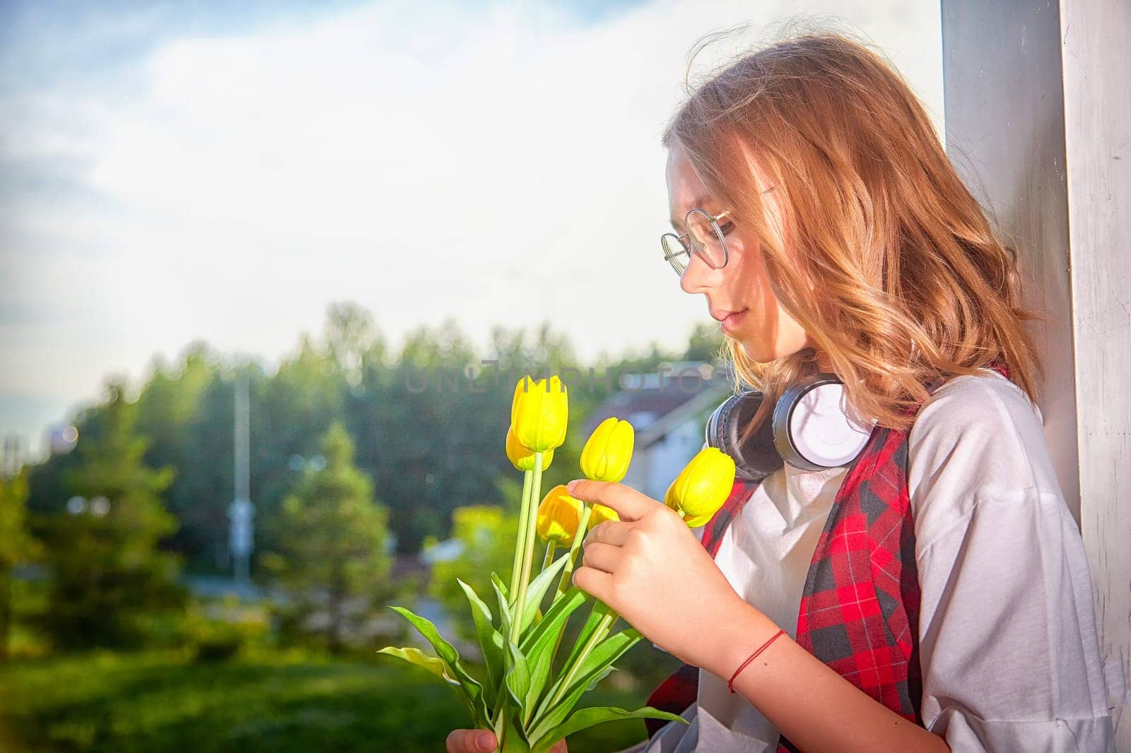 A girl wearing glasses and headphones admires yellow tulips while standing by a window on a bright day.