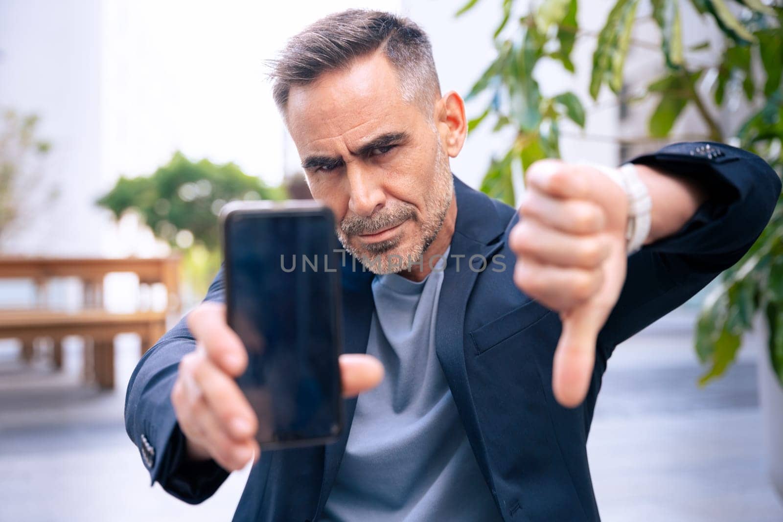 Displeased man in a blazer holding a smartphone and making a thumbs-down but gesture outdoors. He looks frustrated or dissatisfied. The background shows an urban environment with tables and buildings