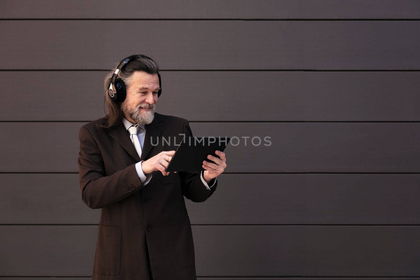 content mature male with beard in formal suit and wireless headphones using tablet while communicating online against gray wall, concept of middle age business man and technology of communication