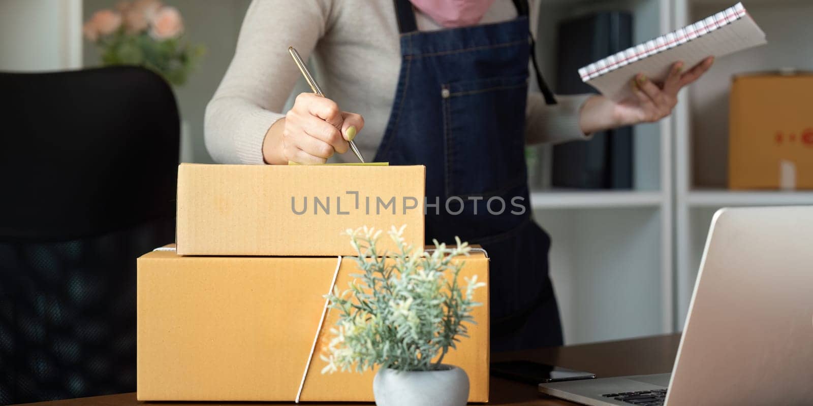 A focused Muslim woman labeling shipping boxes and holding a notebook at her workspace, showcasing her proactive approach to online sales and organization.