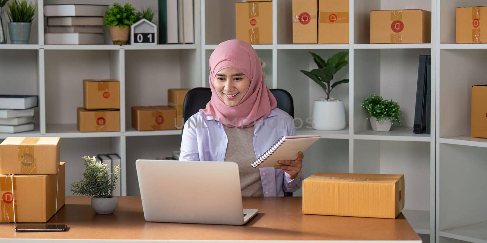 A Muslim woman in a hijab joyfully managing her online sales using a laptop and taking notes in her home office, reflecting her dedication to her business.