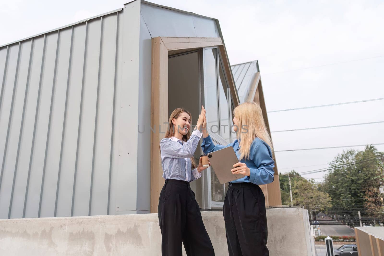 Two businesswomen outdoors giving each other a high-five, celebrating the success of their project while holding a tablet in a modern architectural setting.