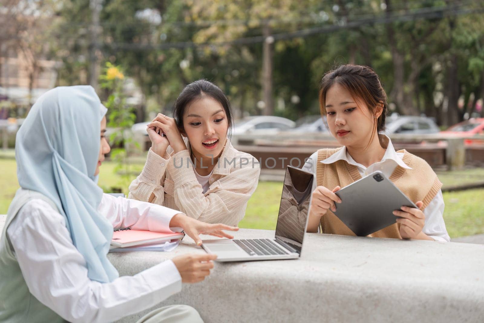 A group of diverse female university students engaging in collaboration, discussing ideas while using a laptop and tablet in a park-like setting.