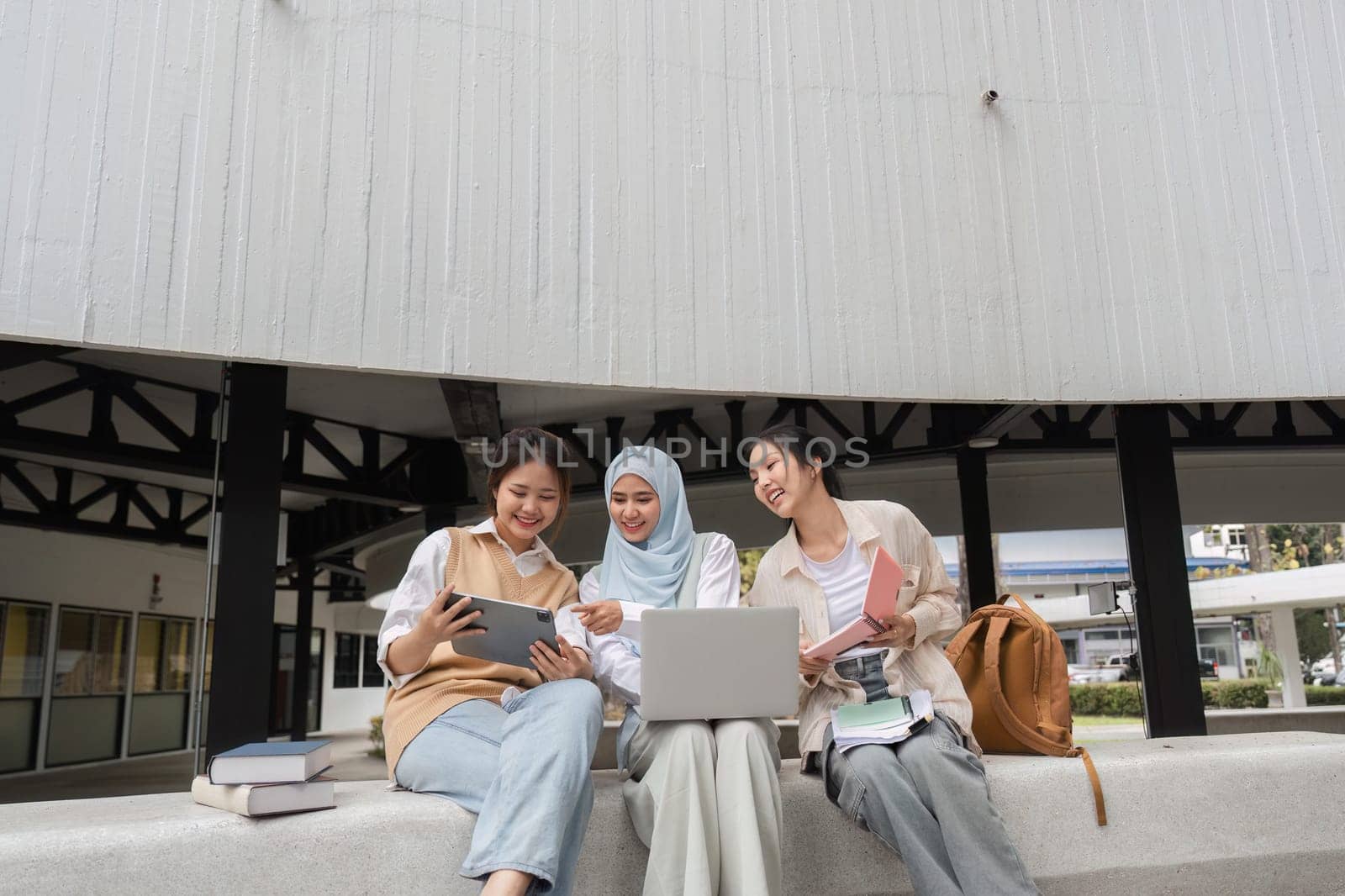 A group of multicultural female students sharing ideas and collaborating using a tablet and laptop, surrounded by books in a university campus environment.