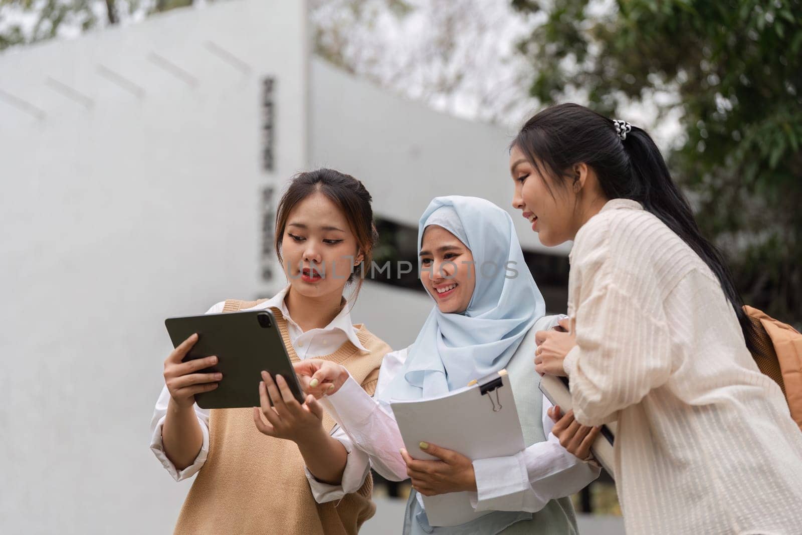 Three diverse female students collaborating around a tablet outdoors, sharing ideas and studying together in a vibrant academic environment.